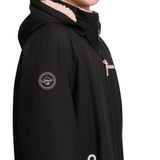 LeMieux AW26 Young Rider HydroShield Robe Varient Black. A close-up side view of a person wearing a black hooded jacket with a light pink fleece-lined hood and light pink zipper accents. A circular logo patch is visible on the left sleeve.