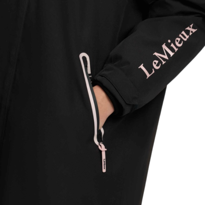 LeMieux AW26 Young Rider HydroShield Robe Varient Black. Close-up of a black jacket sleeve with the brand name 'LeMieux' printed in light pink, featuring a zippered pocket with a black and pink zipper pull.