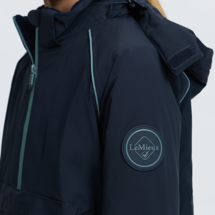 LeMieux AW26 Young Rider Bailey Waterproof Anorak Varient Navy. Close-up of a person wearing a dark navy hooded jacket with light teal piping and a circular LeMieux logo patch on the upper sleeve, against a plain light gray background.