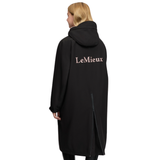 LeMieux AW26 Young Rider HydroShield Robe Varient Black. A woman shown from the back wearing a long black hooded coat with 'LeMieux' printed in a light pink serif font across the upper back, set against a plain white background.