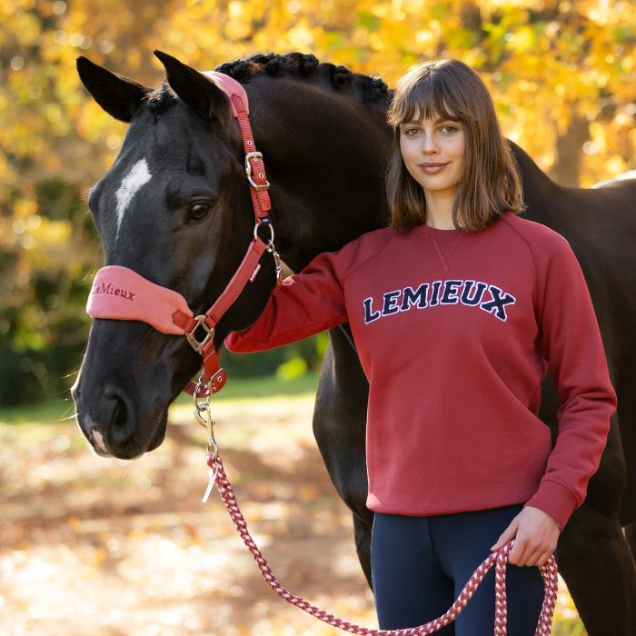 LeMieux AW26 Vida Varsity Crew Neck Sweat Variant Colour Russet.
A woman stands outdoors with a black horse wearing red branded horse gear. The woman wears a red sweatshirt with 'LEMIEUX' printed across the chest in navy and white.