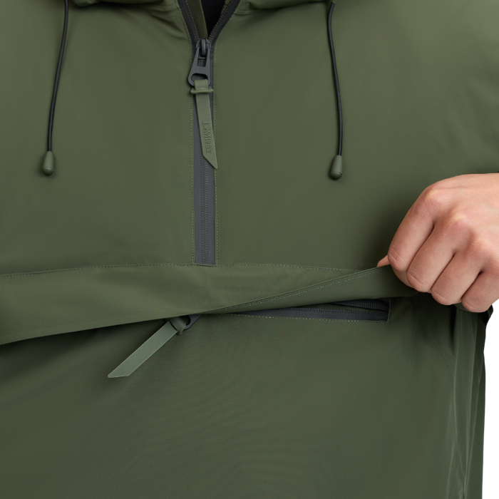 LeMieux AW26 Mens Waterproof Anorak Varient Olive. Close-up of an olive green hooded jacket with a half-zip front, drawstrings, and a hand pulling open a horizontal zippered pocket.