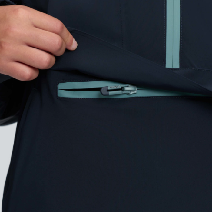 LeMieux AW26 Young Rider Bailey Waterproof Anorak Varient Navy. Close-up of a person's hand lifting the hem of a dark navy jacket to reveal a zippered pocket with a contrasting light teal zipper.