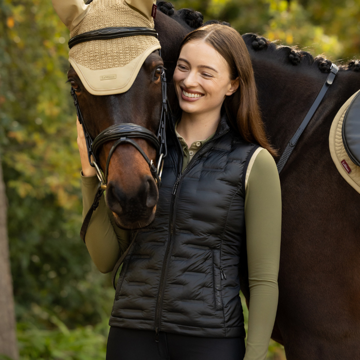 LeMieux AW26 Brooke 3 in 1 Waterproof Hybrid Jacket Varient Black. 
Woman smiling beside a dark brown horse outdoors, wearing a black quilted equestrian vest over an olive green long-sleeve shirt. The horse has tan protective head and neck wraps, and the background is softly blurrey outdoor setting. 