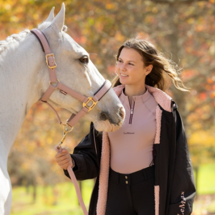 LeMieux AW26 Young Rider HydroShield Robe Varient Black. A woman in equestrian apparel stands beside a white horse outdoors, holding its reins. She wears a pink quarter-zip top, black pants, and a black jacket with pink fleece lining.