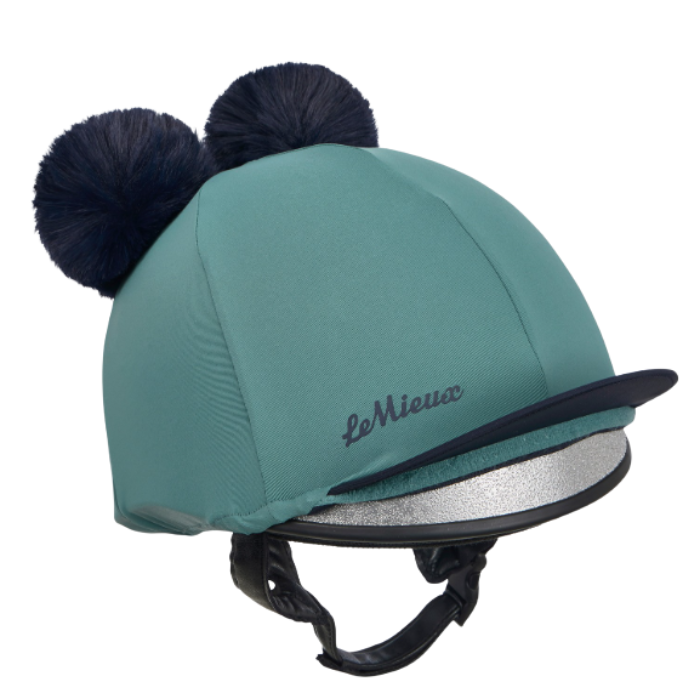 LeMieux AW26 Mini Double Pom Hat Silk Variant Colour Navy/Aloe.
A teal equestrian riding helmet with two dark navy blue pom-poms on top, a black visor, and a silver metallic band around the brim. The brand name 'Le Mieux' is embroidered in black on the side.