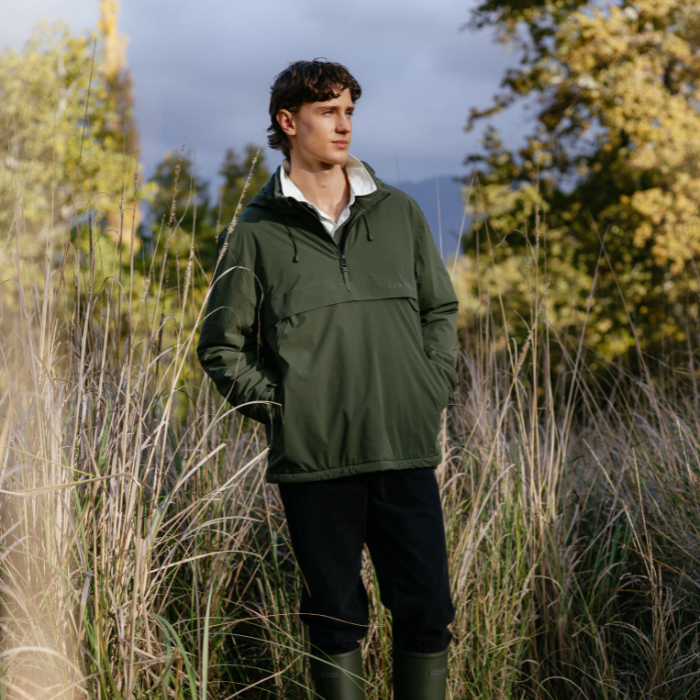 LeMieux AW26 Mens Waterproof Anorak Varient Olive. A man wearing an olive green lightweight pullover jacket with a half-zip and drawstring hood, standing in tall dry grass outdoors with trees and a cloudy sky in the background.