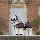 Mini toy pony wearing a plaid rug and matching head collar infront of a brick house.