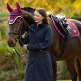 LeMieux AW26 Stormwear Performance Riding Coat Varient Navy. A woman wearing a navy blue riding jacket stands beside a brown horse outdoors. The horse is equipped with a purple fly veil and a black saddle with a purple saddle pad.