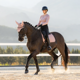 LeMieux AW26 Essence Dressage Square Varient Wild Rose. A rider in a light pink long-sleeve equestrian shirt, black breeches, and a black helmet rides a dark brown horse in a sandy riding arena with a white fence and blurred hills in the background.