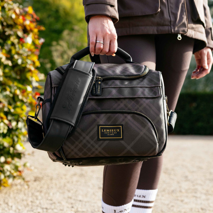 A person outdoors carrying a dark gray and black patterned bag with a black leather handle and shoulder strap. The bag has a gold-tone 'LERMIEUX' logo plaque on the front.