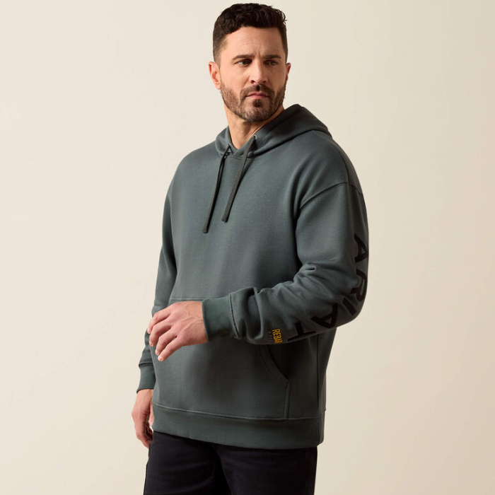 A man wearing a dark slate green pullover hoodie with black drawstrings and black graphic lettering on the sleeve, standing against a plain beige background.