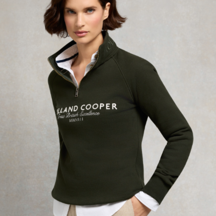 Holland Cooper Sporting Goods Zip Henley variant Racting Green. Woman wearing a green 1/4 zip with Holland Cooper branding on a grey background. 