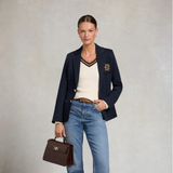 Woman wearing a navy blazer with a logo, holding a brown handbag, on a light gray background
