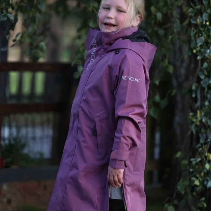 A young girl wearing a long purple REINCOAT raincoat stands outdoors in front of a blurred background of trees and foliage.