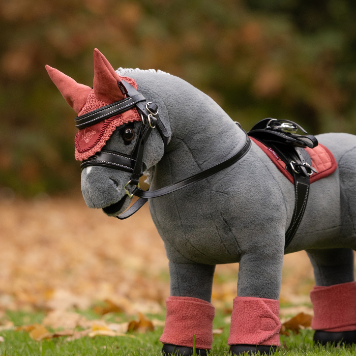 LeMieux AW26 Toy Pony Fly Hood Variant Colour Spiced Earth.
A plush toy horse wearing a spiced earth colour equestrian costume, including a horned bridle, black faux-fur mane, saddle, and leg wraps, standing on grass with a blurred outdoor background