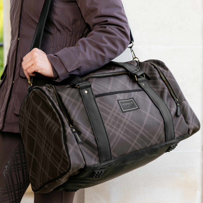 A person wearing a dark plum jacket carries a large brown plaid duffel bag with black trim and a front zip pocket. The brand name 'LeMieux' is visible on the front patch.
