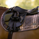 A close-up of an equestrian saddle setup on a horse, featuring a black leather saddle, black girth, and a quilted saddle pad with a brown and beige plaid pattern and a visible Equestrian brand patch.