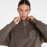 A woman wearing a taupe brown quarter-zip sweatshirt with a silver metallic monogram logo on the left chest, set against a plain white studio background.