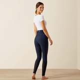 A woman stands barefoot in a studio wearing high-waisted navy equestrian riding breeches with black knee patches and a white short-sleeve top, photographed against a plain beige wall and light wood floor.
