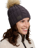 A smiling woman wearing a dark charcoal gray cable-knit beanie with a large tan faux-fur pom-pom and a small dark brand patch on the side, against a plain white background.