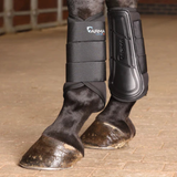 Black Arma Care horse leg boots on a horse's lower legs, shown standing on a tan brick floor in an indoor stable setting.