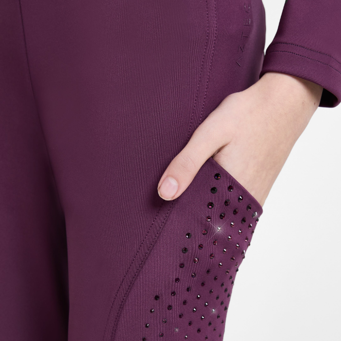 Close-up of a person wearing a fitted athletic legging in a deep plum purple, showing a side pocket with black rhinestone embellishments and a hand reaching into the pocket.