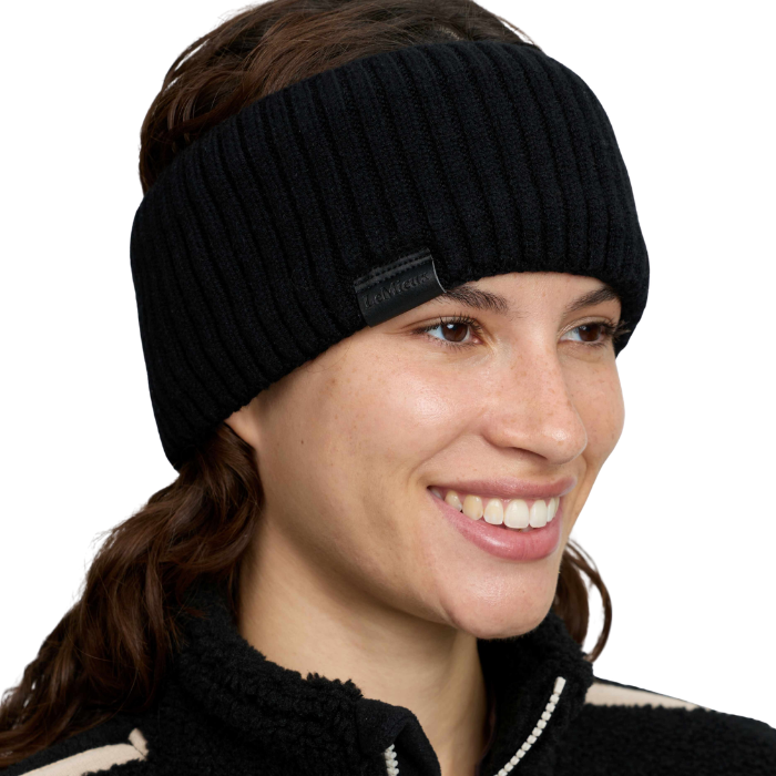 LeMieux AW26 Clara Rib Headband Variant Black. A smiling woman wearing a black ribbed knit headband with a small black brand tab on the side, photographed against a clean white background.