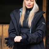 Lady putting on a Black glove whilst wearing a Navy Reincoat infront of a stable background. 