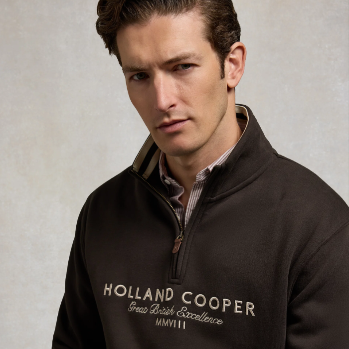 Holland Cooper Heritage Quarter Zip Sweat (Men's) Variant Bournville. Close up of man wearing a 1/4 zip with white Holland Cooper branding and brown zip detaling against a grey background. 