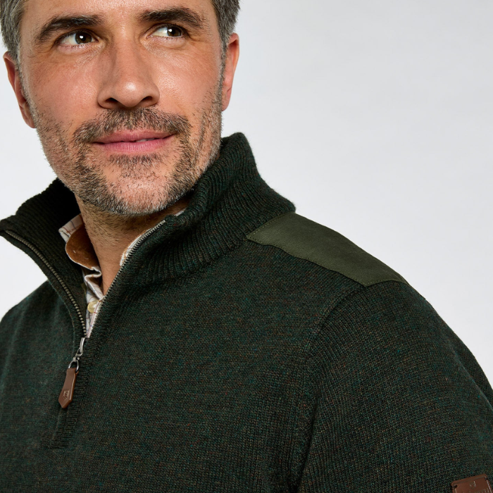 Close-up of a man wearing a dark green quarter-zip knit sweater with olive shoulder panels and brown leather accents, photographed against a light gray studio background.