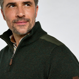Close-up of a man wearing a dark green quarter-zip knit sweater with olive shoulder panels and brown leather accents, photographed against a light gray studio background.
