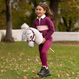 LeMieux AW26 Hobby Horse Coco Variant Colour White.
A young girl stands on a grassy park lawn wearing a matching burgundy and pink equestrian outfit, holding a plush white hobby horse toy.