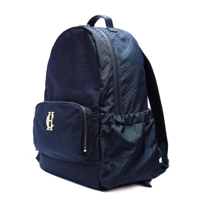 Holland Cooper Burghley Backpack Variant Ink Navy Monogram. A navy blue backpack with a subtle geometric pattern, featuring a front zippered pocket with a gold-tone 'HC' logo and a side mesh pocket, shown on a white background.
