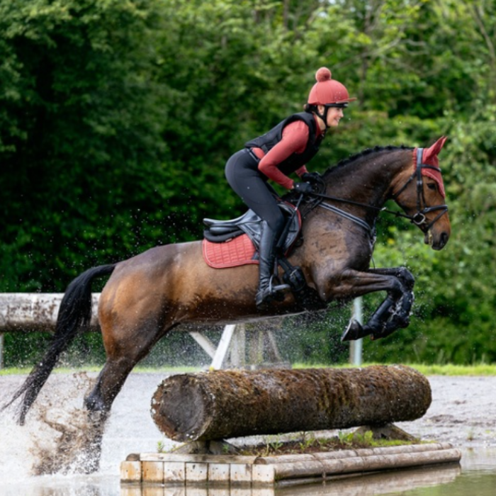 LeMieux AW26 Suede Close Contact Square Varient Russet. A rider in red and black equestrian attire jumps a dark brown horse over a log obstacle in an outdoor water course, with green trees in the background.