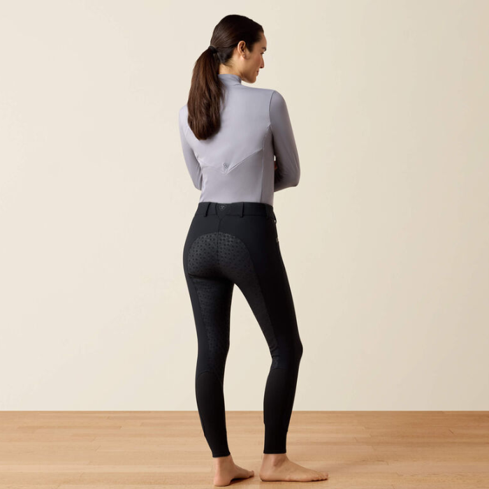 A woman stands barefoot in profile, wearing black equestrian riding breeches with a textured pattern and a light gray long-sleeve top, set against a plain beige wall and light wood floor.