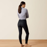 A woman stands barefoot in profile, wearing black equestrian riding breeches with a textured pattern and a light gray long-sleeve top, set against a plain beige wall and light wood floor.