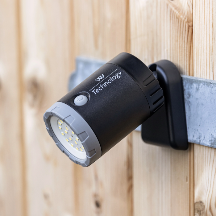 A black outdoor spotlight with a gray front trim and white LED lenses, mounted on a gray metal bracket against a light wood plank wall. The spotlight has 'W Technology' printed on the side.