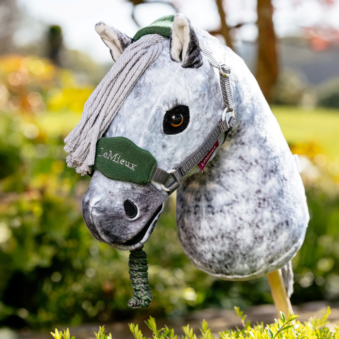 LeMieux AW26 Hobby Horse Vogue Headcollar & Leadrope Variant Colour Hunter Green. 
A plush toy horse head mounted on a stick, with a white body, gray faux-fur mane, and hunter green colour halter, photographed outdoors against a blurred garden background.