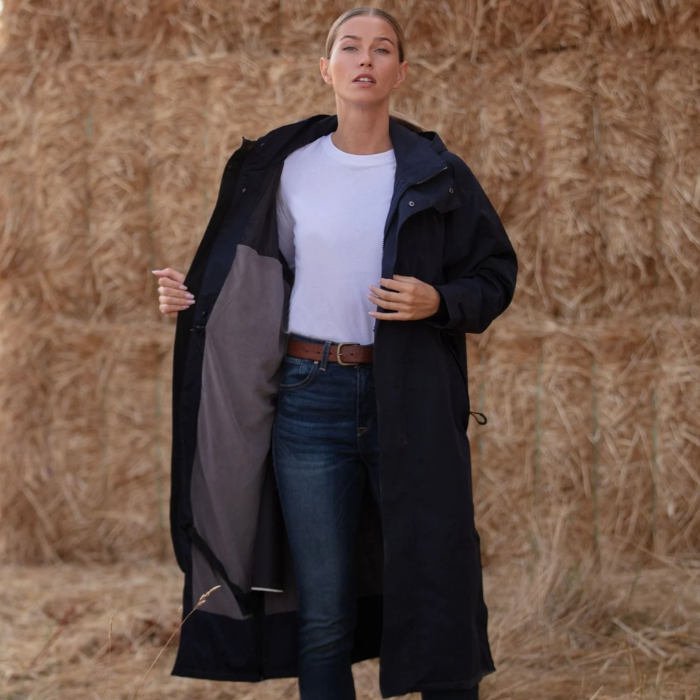 A woman stands outdoors in front of a hay bale wearing a long black hooded coat over a white top and blue jeans, holding the coat open to show the gray interior lining.