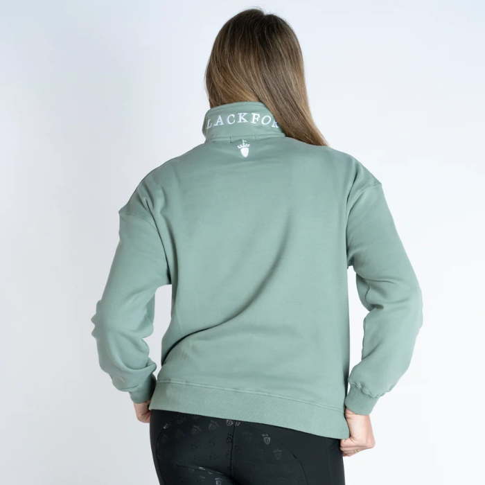 Blackfort Equestrian Ella Quarter Zip Variant Sage. A woman viewed from the back wearing a sage green quarter-zip sweatshirt with 'BLACKFORT' printed in white on the collar, paired with black pants, against a plain white studio background.