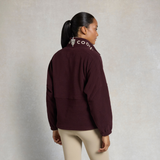 Holland Cooper Cranborne Over Head Fleece Variant Mulberry. A woman shown from the back wearing a dark maroon fleece jacket with a high collar featuring white text and a shield logo, paired with beige leggings, against a textured light gray background.