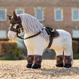 A plush toy horse styled like a dapple gray horse, wearing a gray shawl mane, black bridle, brown saddle, and brown plaid leg wraps, standing on a gravel path with a blurred brick building in the background.