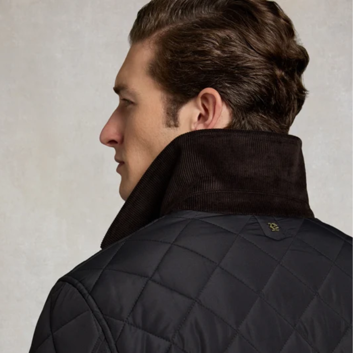A close-up side profile of a man wearing a dark quilted jacket with a high corduroy collar, photographed against a light textured studio background.