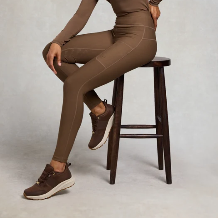A model wearing a matching brown activewear set consisting of a long-sleeve top and high-waisted leggings, sitting on a dark wooden stool. The model is also wearing brown athletic sneakers with white soles, set against a neutral light gray studio background.