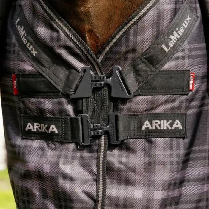 Close-up of a horse blanket with a gray and black plaid pattern, featuring black straps with white 'LeMieux' and 'ARIKA' branding and t-bar buckle closures.