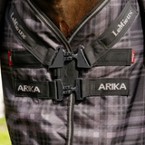 Close-up of a horse blanket with a gray and black plaid pattern, featuring black straps with white 'LeMieux' and 'ARIKA' branding and t-bar buckle closures.