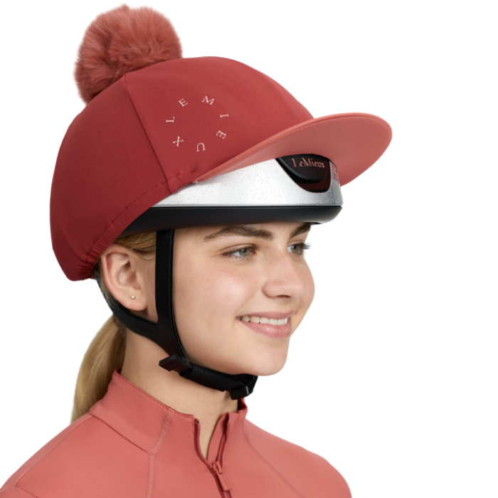 LeMieux AW26 Classique Pom Hat Silk Variant Colour Russet
A woman in profile wearing a equestrian riding helmet with a russet red cover and matching pom-pom and visor, featuring the brand name 'Le Mieux' printed on the side, set against a solid white background.