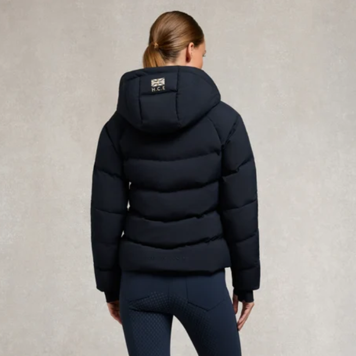 Holland Cooper Chiltern Yard Jacket Variant Ink Navy. A woman viewed from the back wearing a dark navy hooded puffer jacket with a small white logo patch on the back of the hood, paired with dark riding tights against a textured light gray studio background.