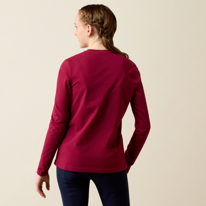Ariat Yth Painted Dream Long Sleeve T-Shirt Varient Beet Red. Girl wearing a red long sleeve top and black trousers from the back on a beige background.